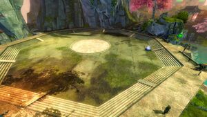 An image of the arena in the Isle of Reflection guild hall, showing a flat octagonal space with shallow steps on each side, a circle marked on the floor in the center, a teleporter on the right side and the arena proprieter NPC in the lower right corner.
